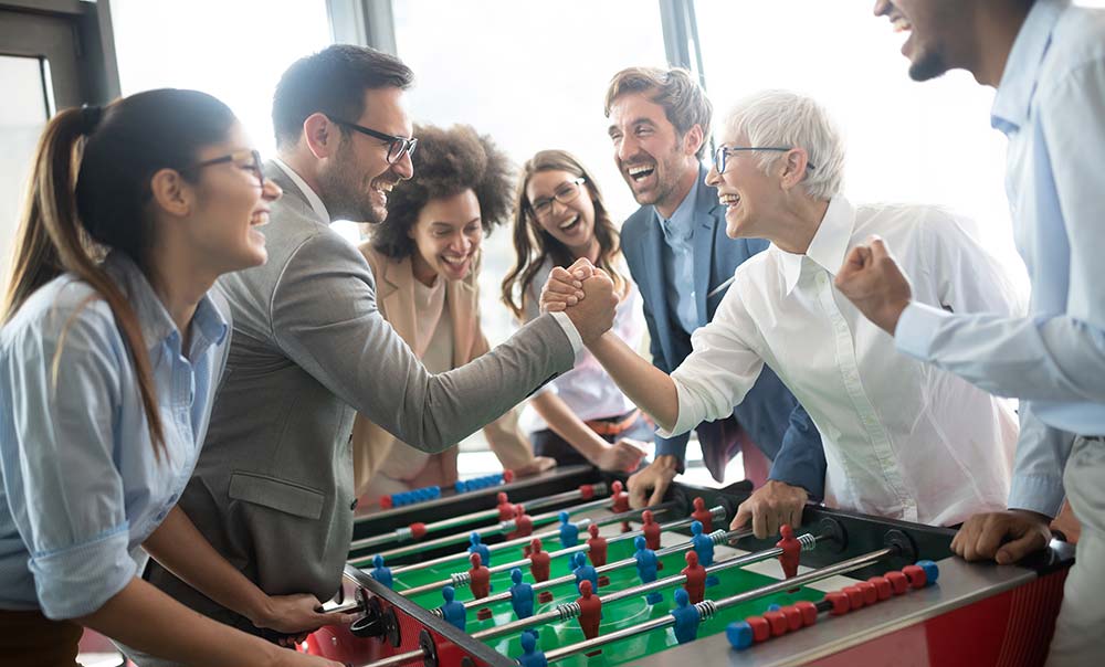 Das dynamische und diverse Team von PB TGA lacht und zeigt beim Tischkickern im Büro den starken Zusammenhalt und eine positive Arbeitsatmosphäre